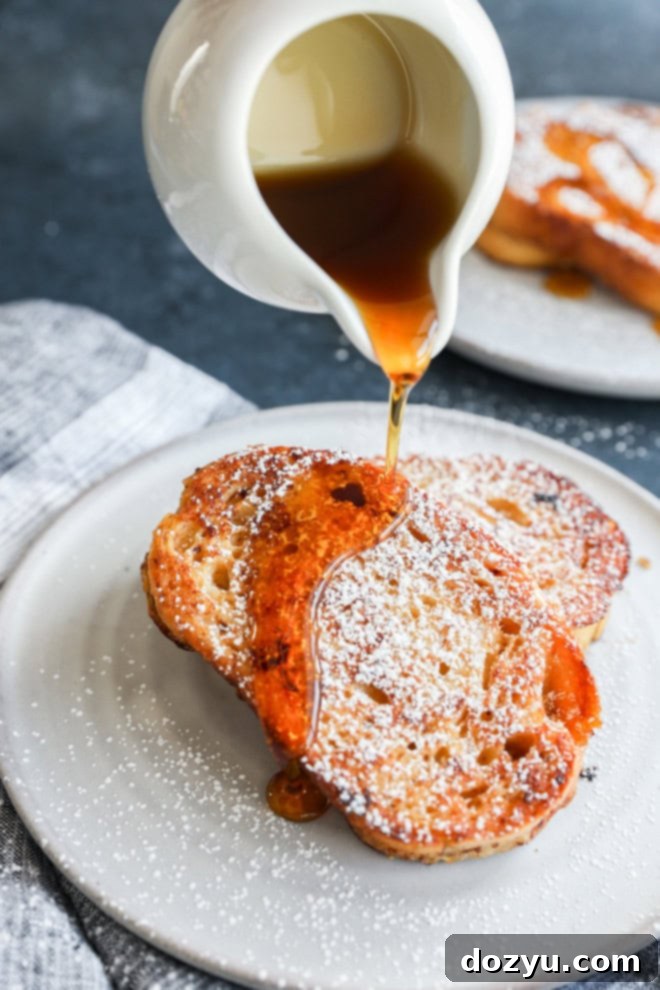 A close-up shot of warm, golden sourdough French toast slices stacked on a plate, being drizzled generously with maple syrup, showcasing its tempting glaze and inviting texture.