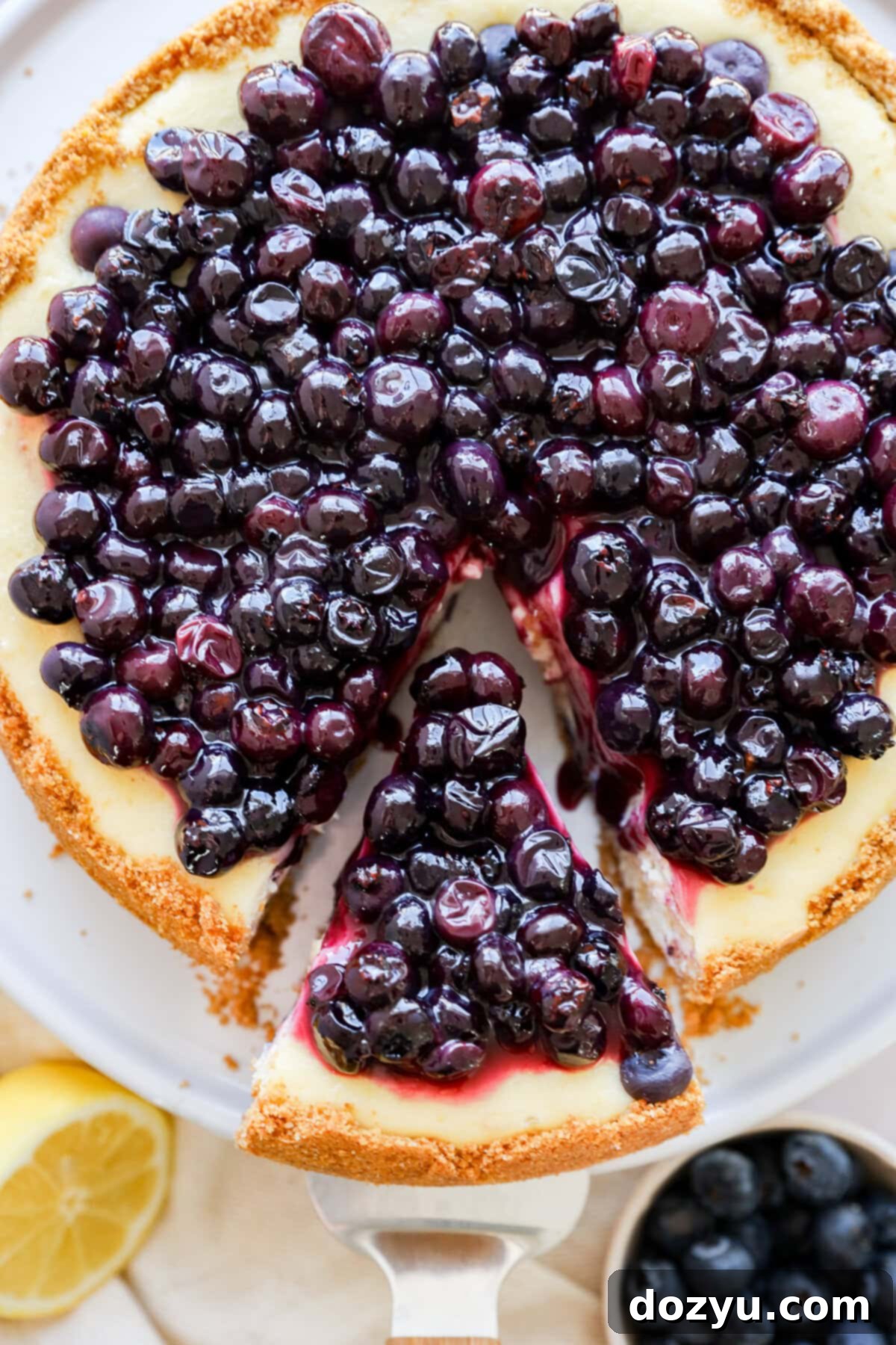 A glorious slice being taken out of a Lemon Blueberry Cheesecake, revealing its creamy texture and fresh blueberries, served on an elegant cake stand.