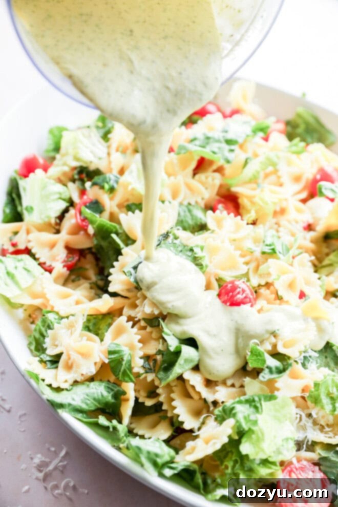 Sun-Kissed Caprese Bow Tie Salad 7 Someone pouring creamy basil dressing over a vibrant Caprese bow tie pasta salad in a large bowl, showcasing the rich texture of the sauce.