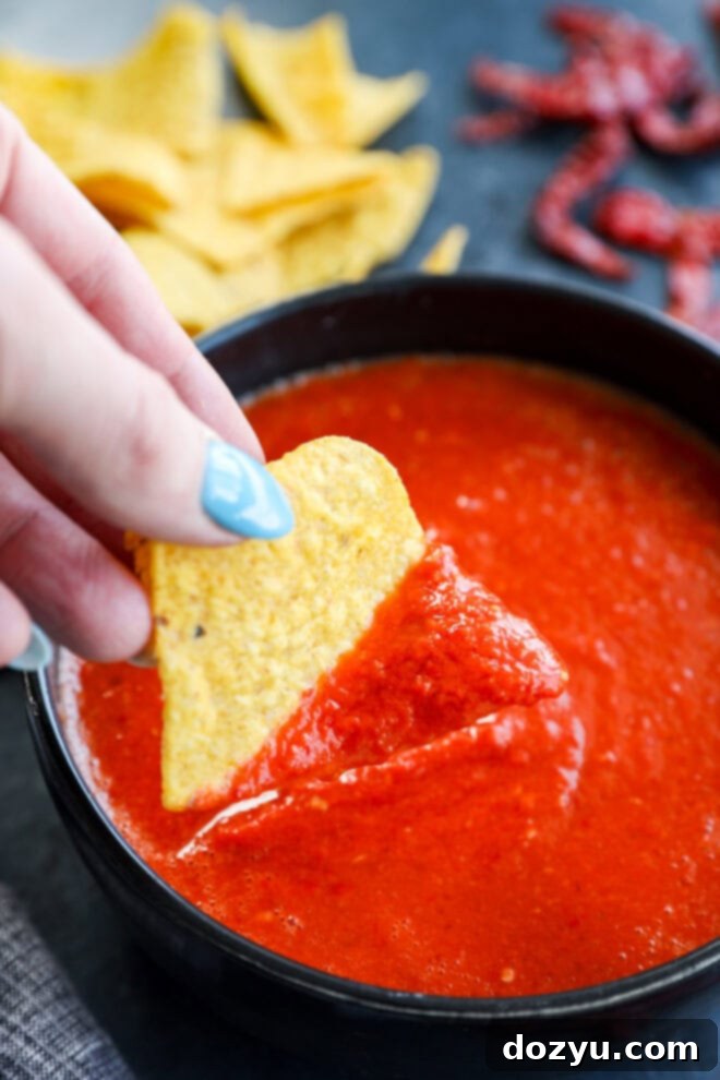 A hand dipping a tortilla chip into a bowl of vibrant red chile de árbol salsa.