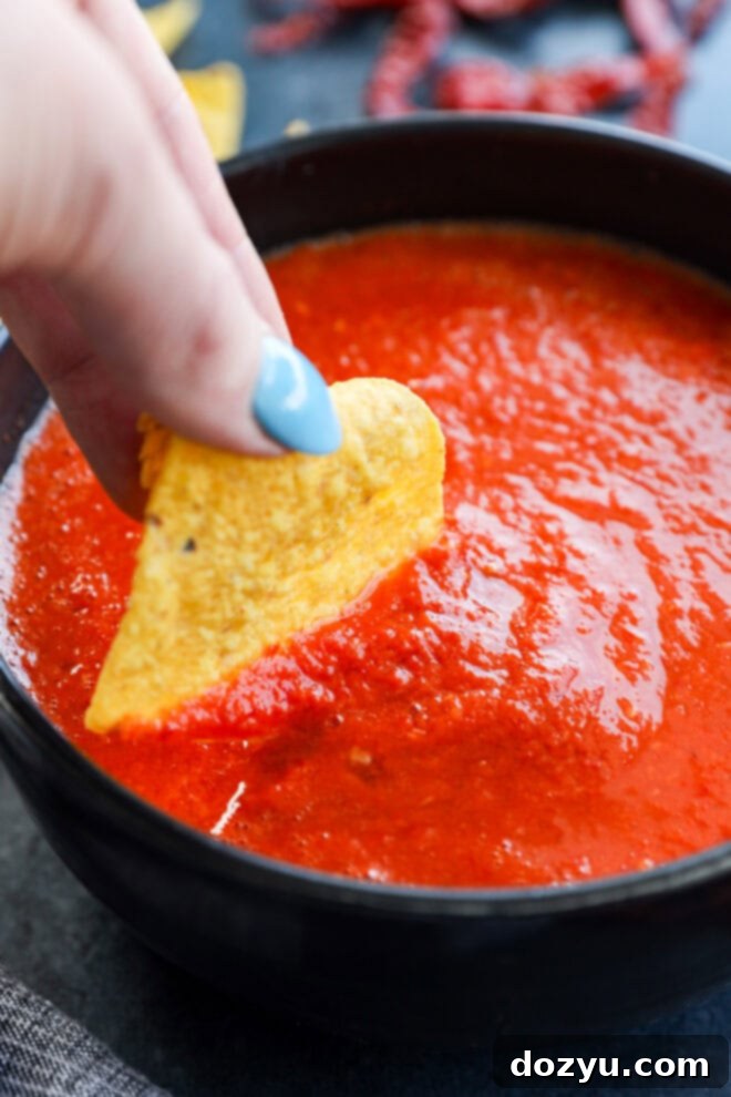 A hand dipping a crispy tortilla chip into a small bowl of spicy red chile de árbol salsa.
