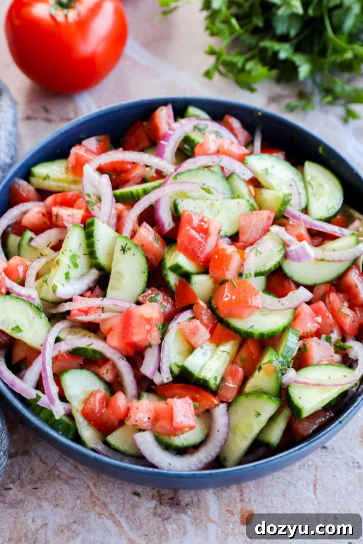 Summer Cucumber Tomato Salad 9 Fresh vegetables and herbs artfully arranged in the background of a blue bowl filled with a vibrant summer side dish, highlighting the freshness of the ingredients.