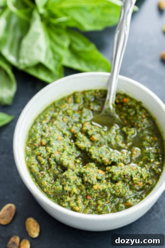 Vibrant Pistachio Pesto 2 A white bowl brimming with vibrant green pistachio pesto, a spoon resting within, flanked by shelled pistachios and fresh basil leaves in the background.