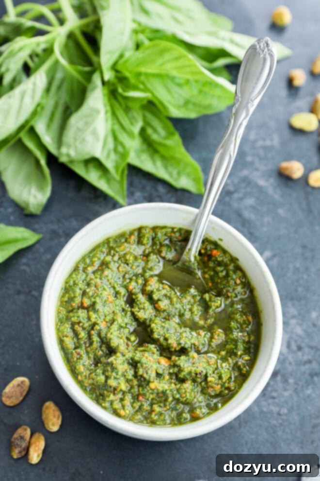 Vibrant Pistachio Pesto 6 A generous serving of vibrant basil sauce in a white pottery bowl, with a spoon resting in it, accompanied by a fresh bunch of basil leaves on the side.