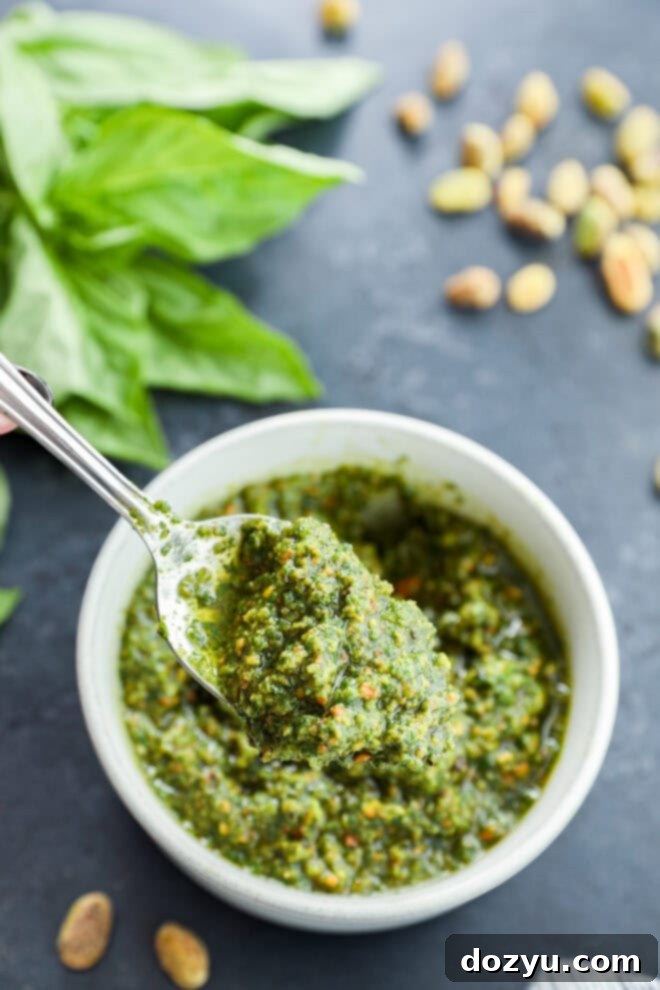 Vibrant Pistachio Pesto 9 A spoon lifting out a vibrant green scoop of pistachio pesto from a white bowl, with shelled pistachios and a bunch of fresh basil arranged artfully in the background.