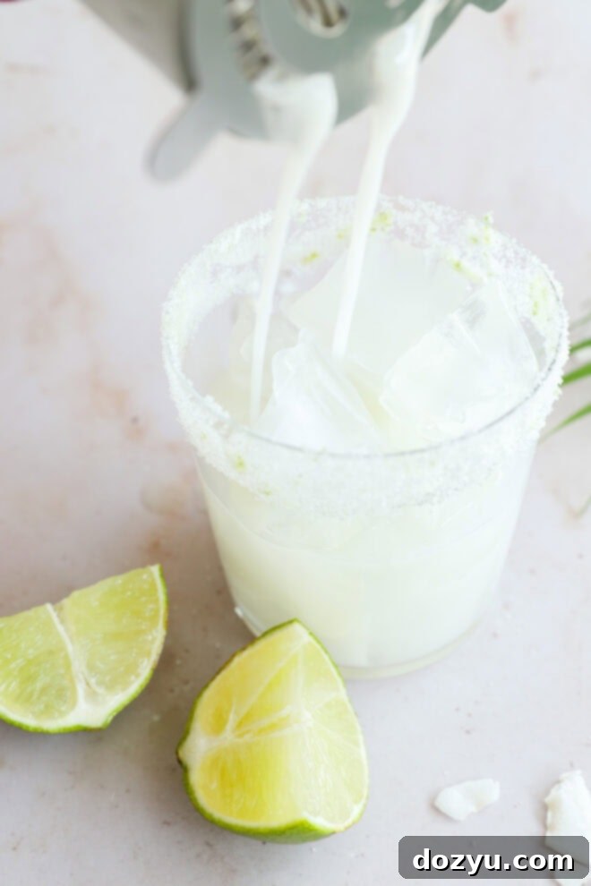 A glass rimmed with sugar and filled with ice cubes is being topped with a milky white Coconut Mojito. Two lime wedges are placed on the surface next to the glass, highlighting the drink's fresh ingredients.