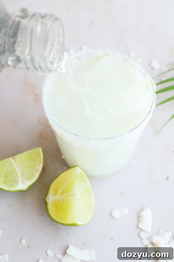 A glass with a sugar-lime rim and ice cubes is being filled with a clear club soda, surrounded by lime wedges and coconut flakes—perfect for serving a refreshing Coconut Mojito on a light surface.