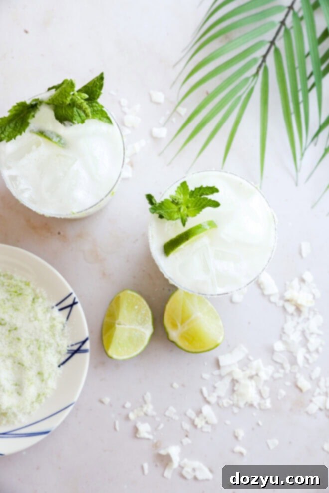 Two glasses of iced Coconut Mojito sit on a white surface with mint and lime garnish, next to lime halves, coconut flakes, a plate with a lime-salt mixture, and a palm leaf, creating a fresh and inviting scene.