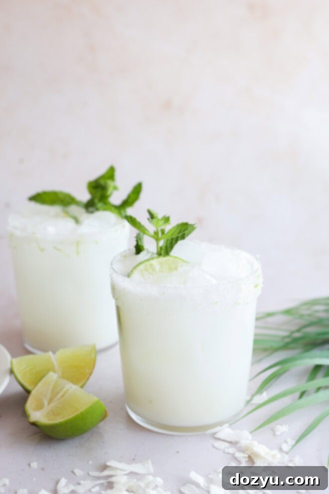 Two glasses of creamy Coconut Mojito with ice, garnished with mint leaves and lime wedges, sit on a light surface. Lime wedges and coconut flakes are scattered nearby. The background is neutral and minimalistic, emphasizing the refreshing drinks.