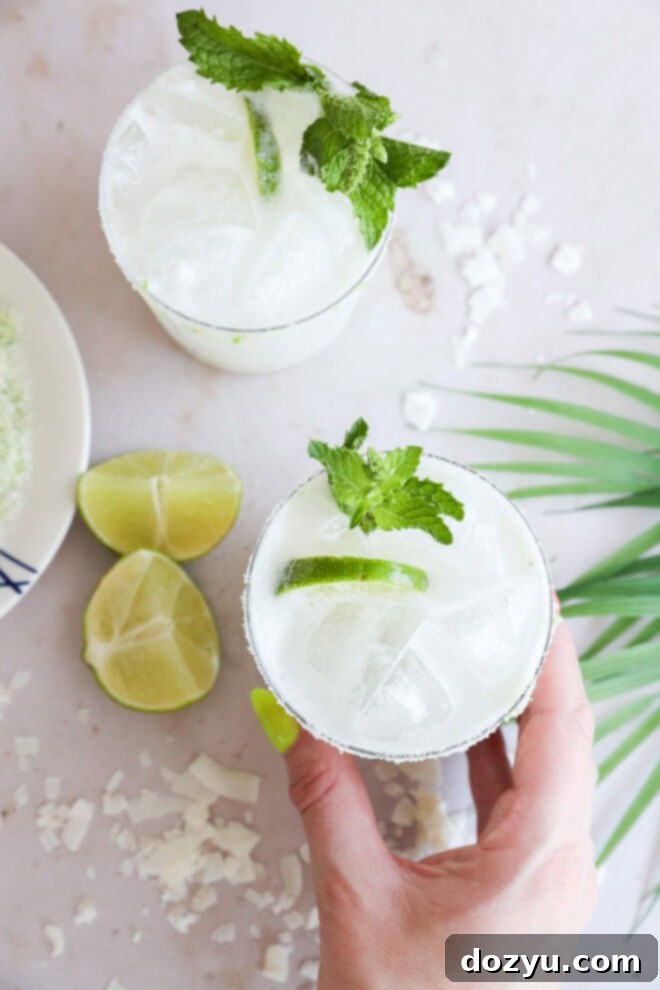 A hand holds a glass of cold, white Coconut Mojito garnished with lime slices and mint. Another similar drink, lime wedges, and coconut flakes are on a light surface nearby, along with a palm leaf decor, suggesting a tropical setting.