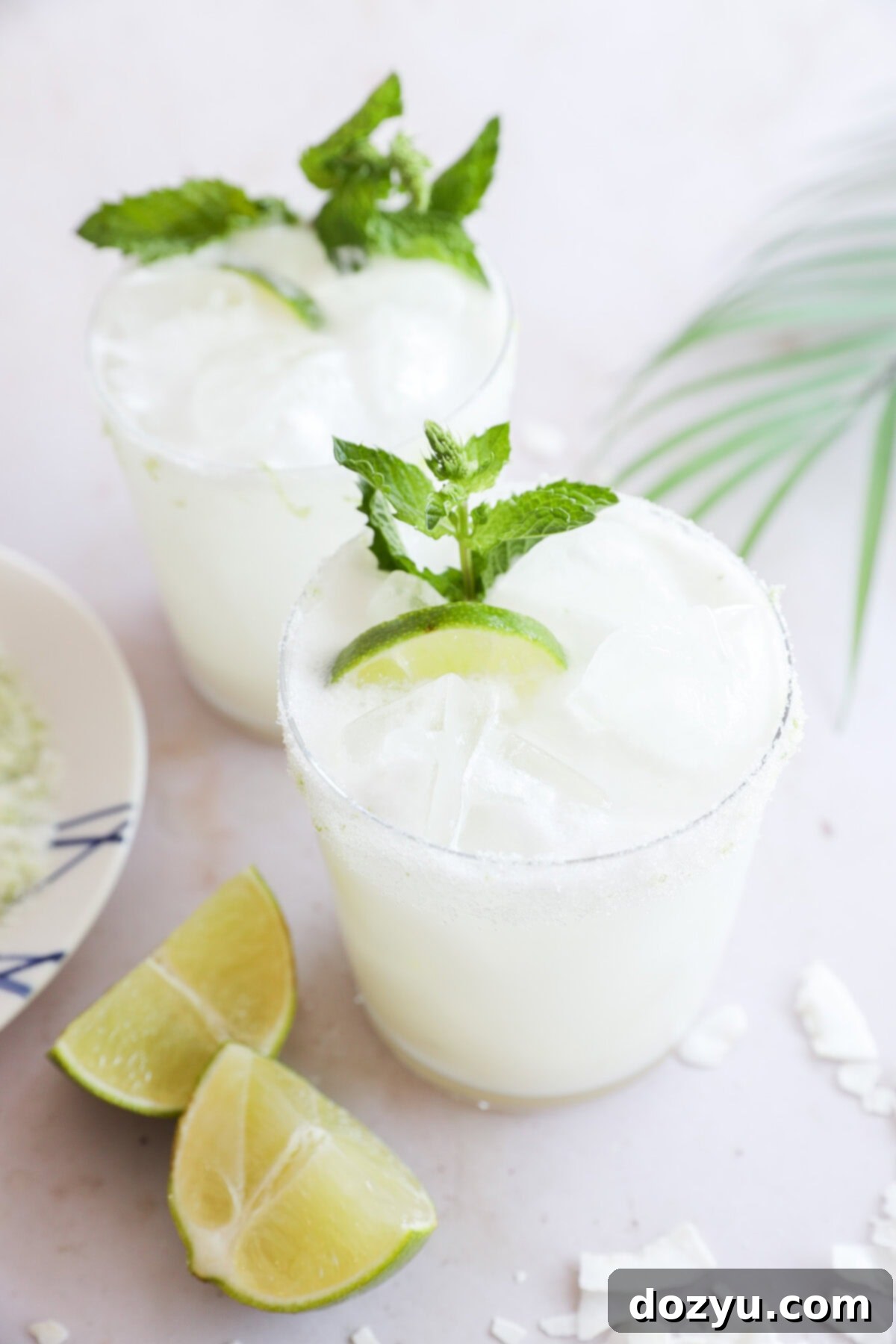 Two glasses of a white, frothy drink with ice, are garnished with lime wedges and fresh mint. Lime wedges and coconut flakes sit on the white surface nearby, with a palm leaf in the background, creating a tropical setting.