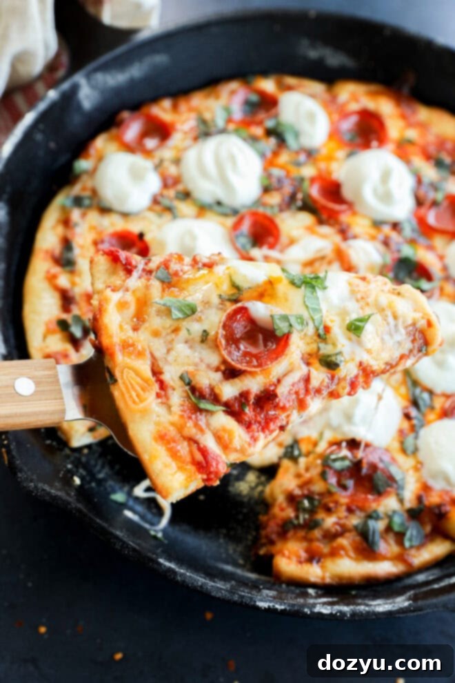 A close-up of a spicy pizza slice being lifted from a cast iron skillet, topped with melted cheese, fresh basil, and dollops of creamy white sauce. The rest of the pizza is visible in the skillet.