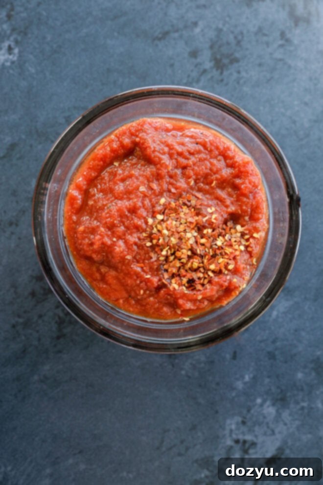 A glass bowl filled with thick red tomato sauce, perfect for spicy pizza, is topped with a sprinkle of crushed red pepper flakes and sits on a dark gray surface.