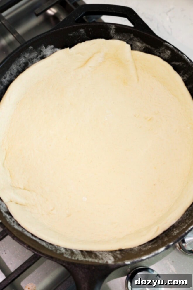 Unbaked pizza dough pressed evenly into a black cast iron skillet, resting on a stovetop with metal burner knobs visible.