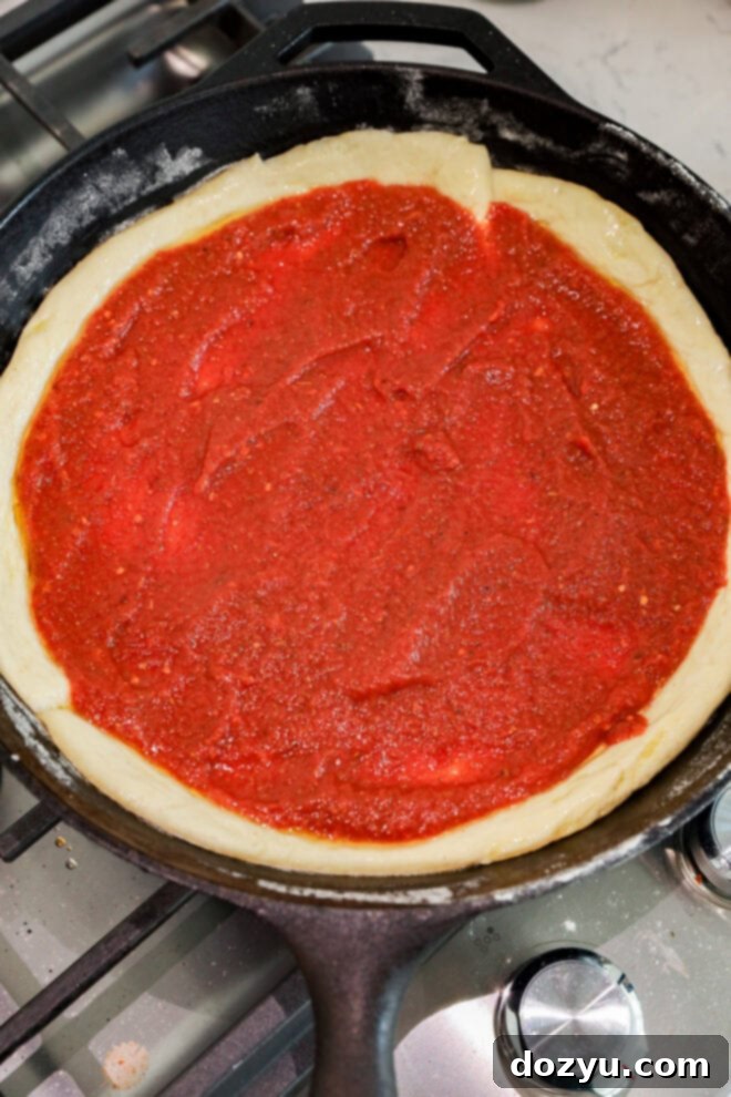 A cast iron skillet on a stove contains spicy pizza dough topped with a layer of tomato sauce, ready for additional toppings and baking.