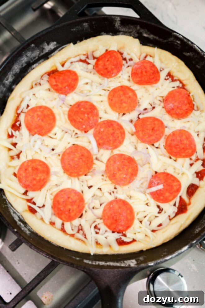 A cast iron skillet on a stove holds an uncooked spicy pizza, topped with shredded mozzarella cheese and pepperoni slices layered over zesty tomato sauce.