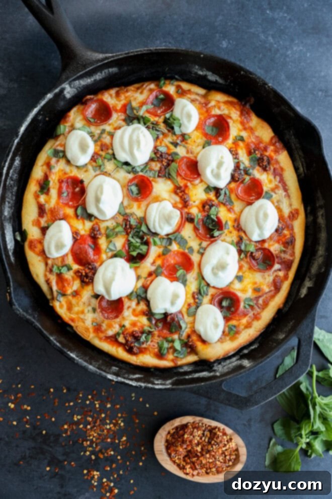 A freshly baked spicy pizza topped with pepperoni, melted cheese, basil, and dollops of ricotta in a black cast-iron skillet, with red pepper flakes and basil leaves on the dark surface nearby.