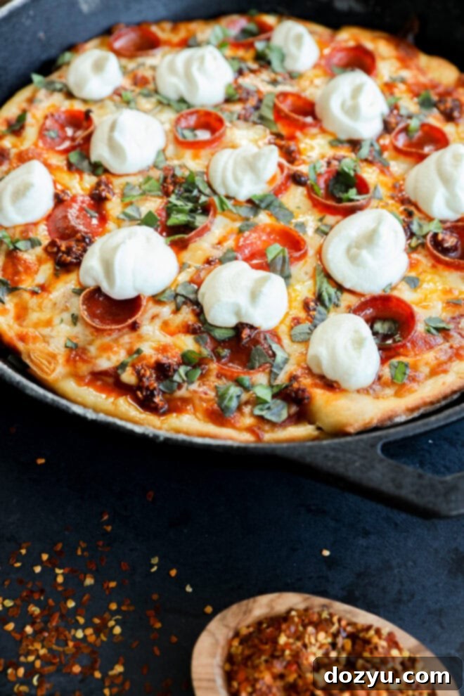 A spicy pizza skillet topped with pepperoni, melted cheese, creamy ricotta, fresh basil, and a mix of spices, served with a wooden spoon of crushed red pepper flakes nearby.