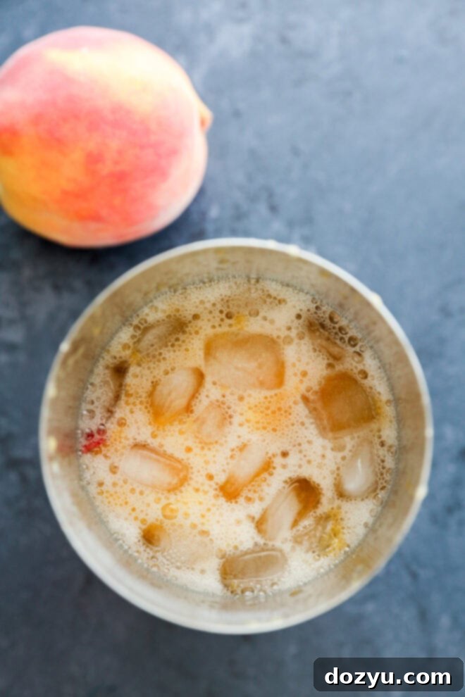 A metal cup filled with an ice and peach-colored bubbly drink sits on a dark surface next to a whole peach.