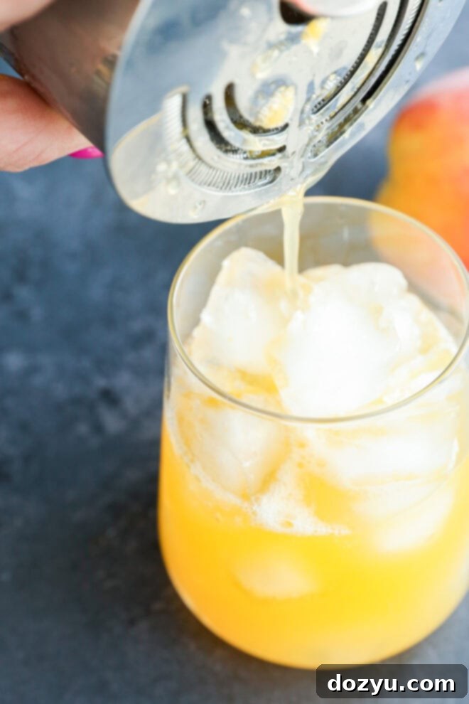 A close-up of a hand pouring a yellow-orange drink over ice cubes in a glass using a cocktail shaker and strainer; a peach is visible in the background on a dark surface.