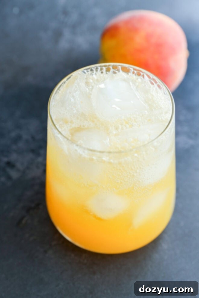 A glass filled with ice and a light orange, fizzy drink sits on a dark surface, with a whole peach blurred in the background.