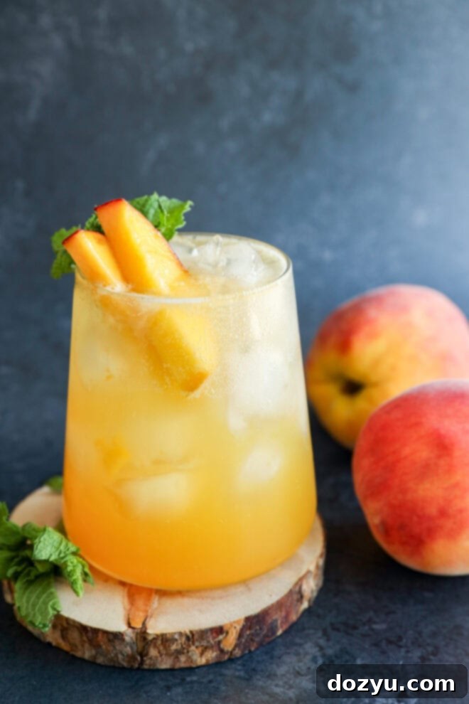 A glass of iced peach drink garnished with peach slices and mint sits on a wooden coaster. Two whole peaches and a sprig of mint are nearby, all set against a dark textured background.