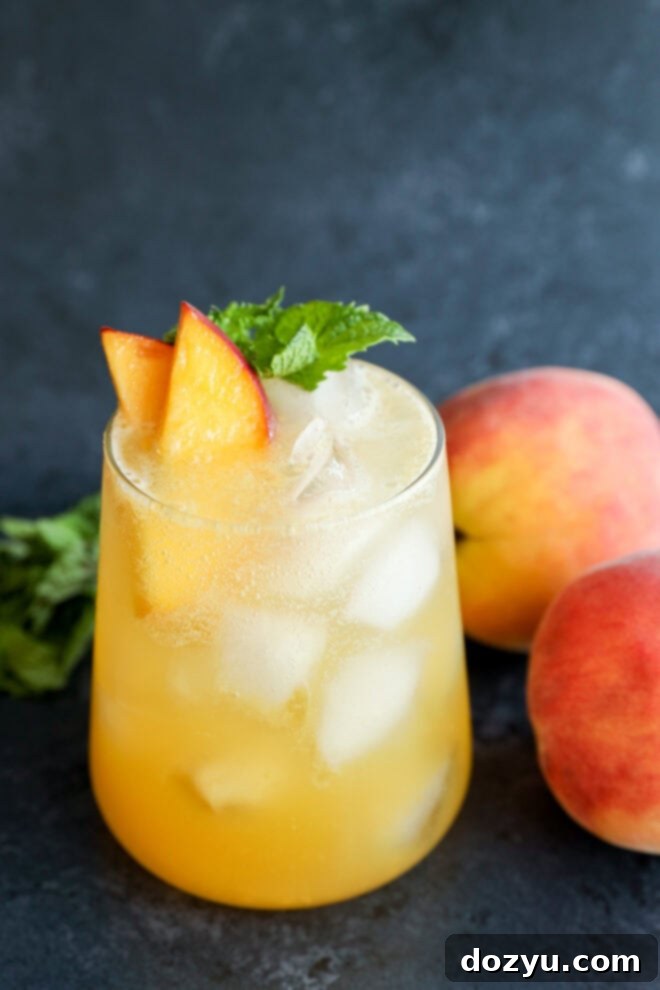 A glass filled with an iced peach-colored drink, garnished with peach slices and mint, sits on a dark surface next to two whole peaches and some fresh mint leaves.