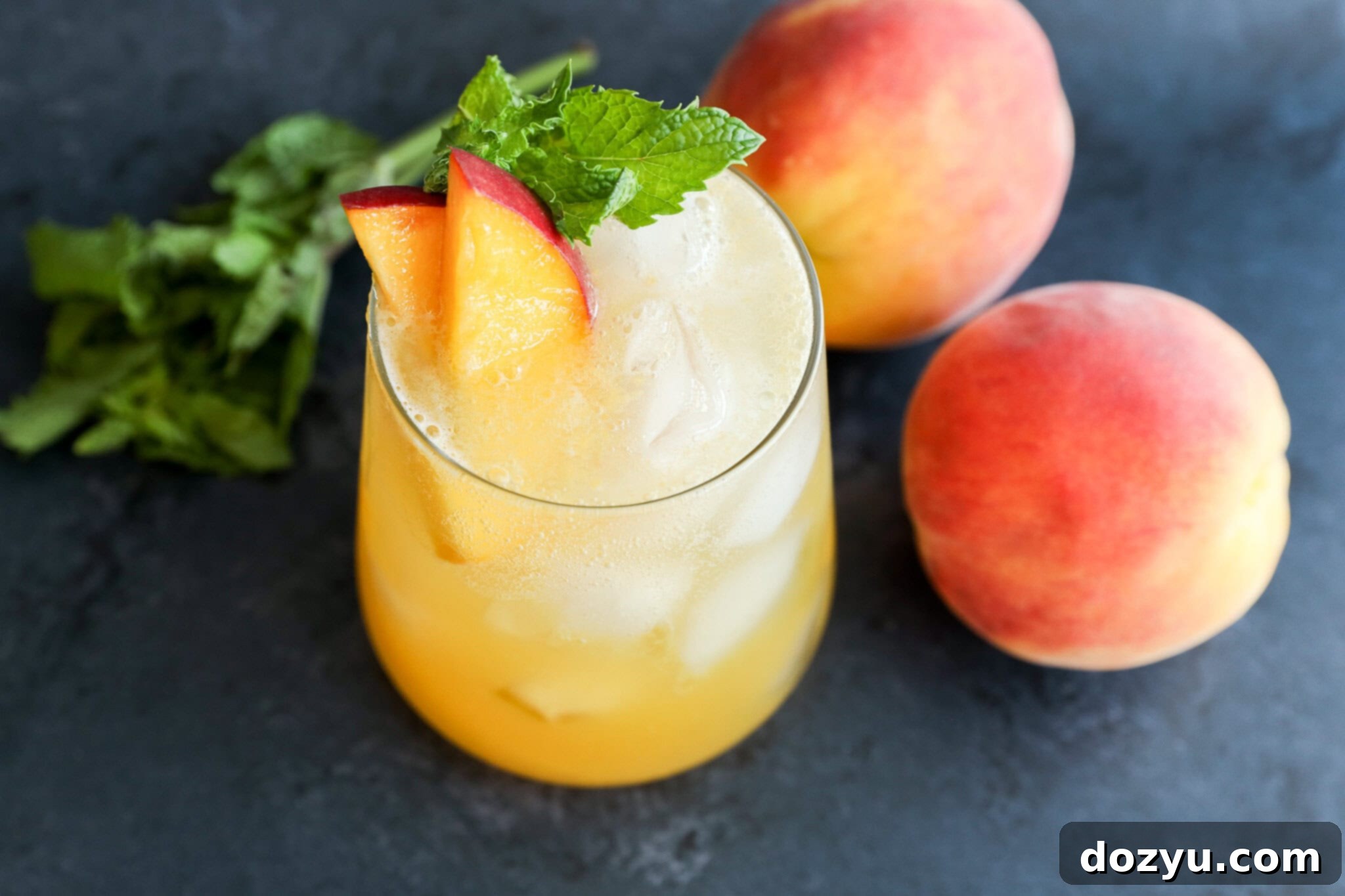 A glass of iced peach drink garnished with peach slices and mint leaves, placed next to two whole peaches and a small bunch of mint on a dark surface.