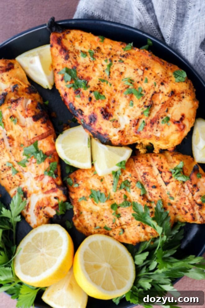 Three grilled, seasoned chicken breasts garnished with fresh parsley are arranged on a black plate with lemon wedges and parsley sprigs. A gray napkin is partially visible in the background.