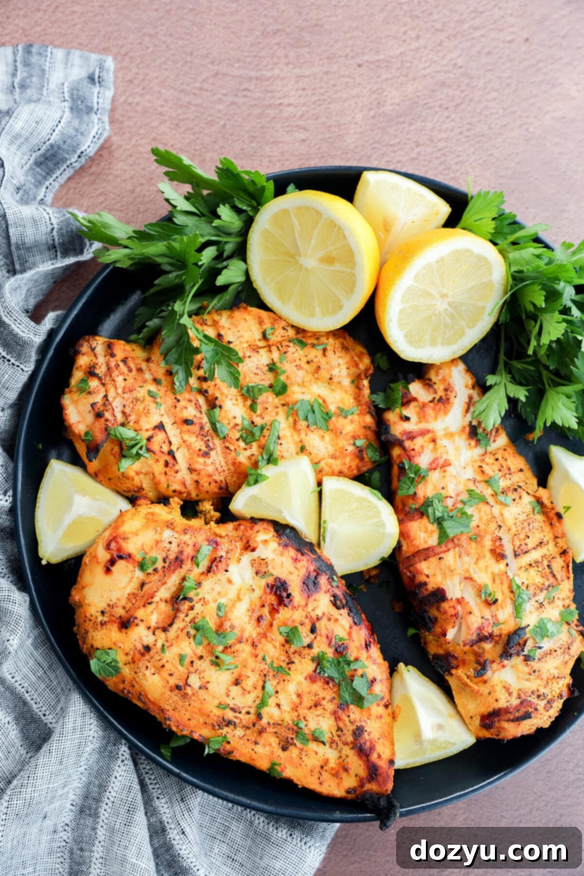 A black plate with three grilled chicken breasts garnished with chopped parsley and surrounded by fresh parsley sprigs and lemon wedges, set on a textured surface with a striped cloth.