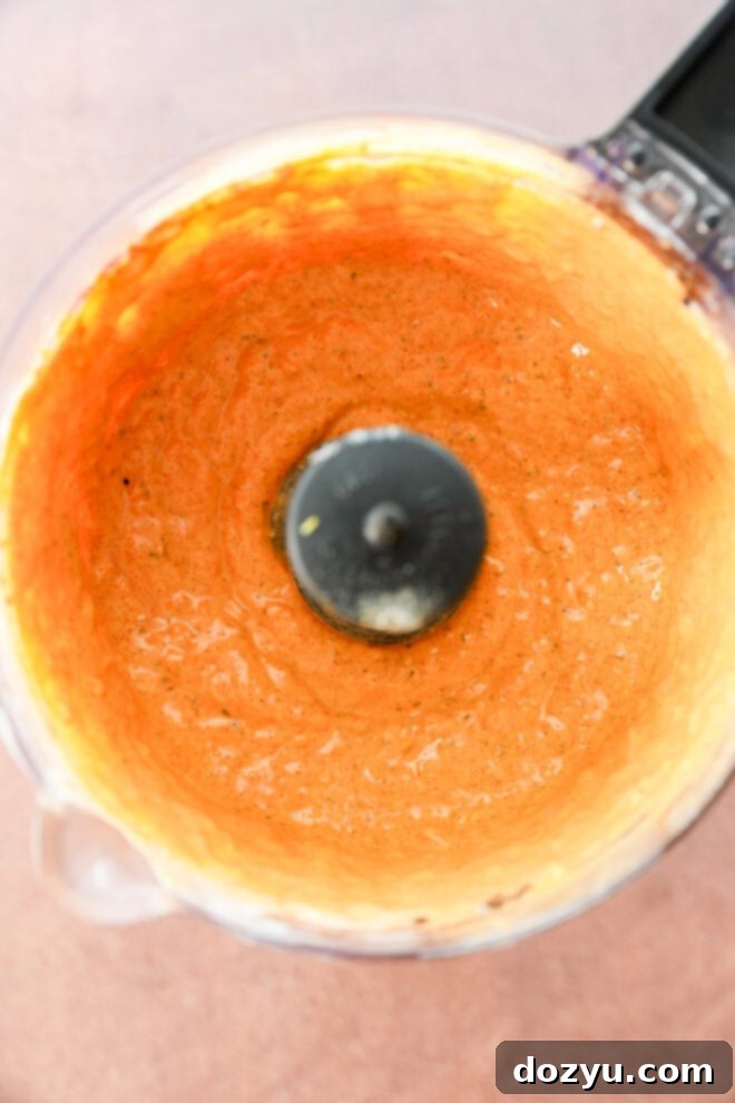 A food processor bowl containing smooth, orange-colored sauce or dip, viewed from above. The sides of the bowl are coated with the mixture, and the black blade is visible in the center.