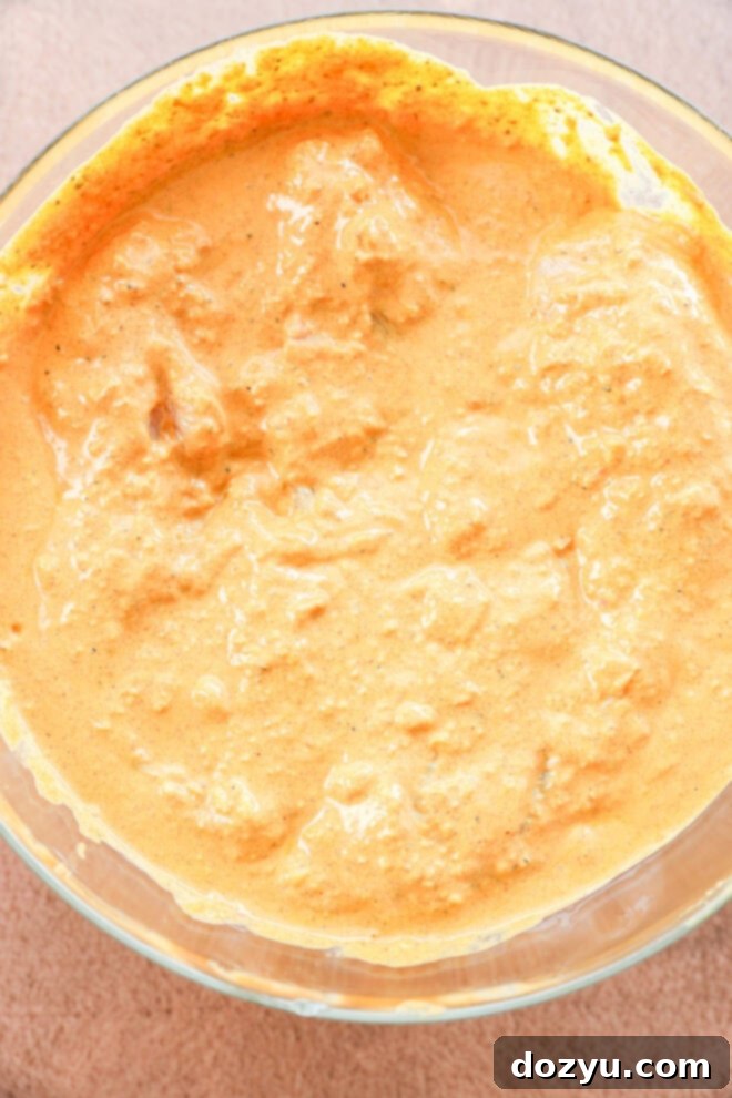A close-up of a glass bowl filled with a thick, creamy orange-colored marinade with visible spices and small chunks, set on a light textured surface.