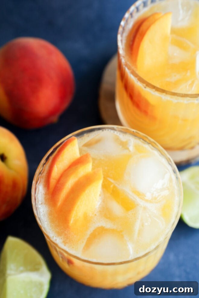 Two glasses of iced peach margarita garnished with fresh peach slices, surrounded by whole peaches and lime wedges on a dark blue surface.