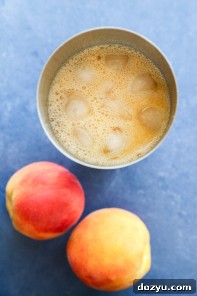 A cocktail shaker filled with an orange, frothy peach margarita and ice cubes is placed on a blue surface next to two whole ripe peaches.