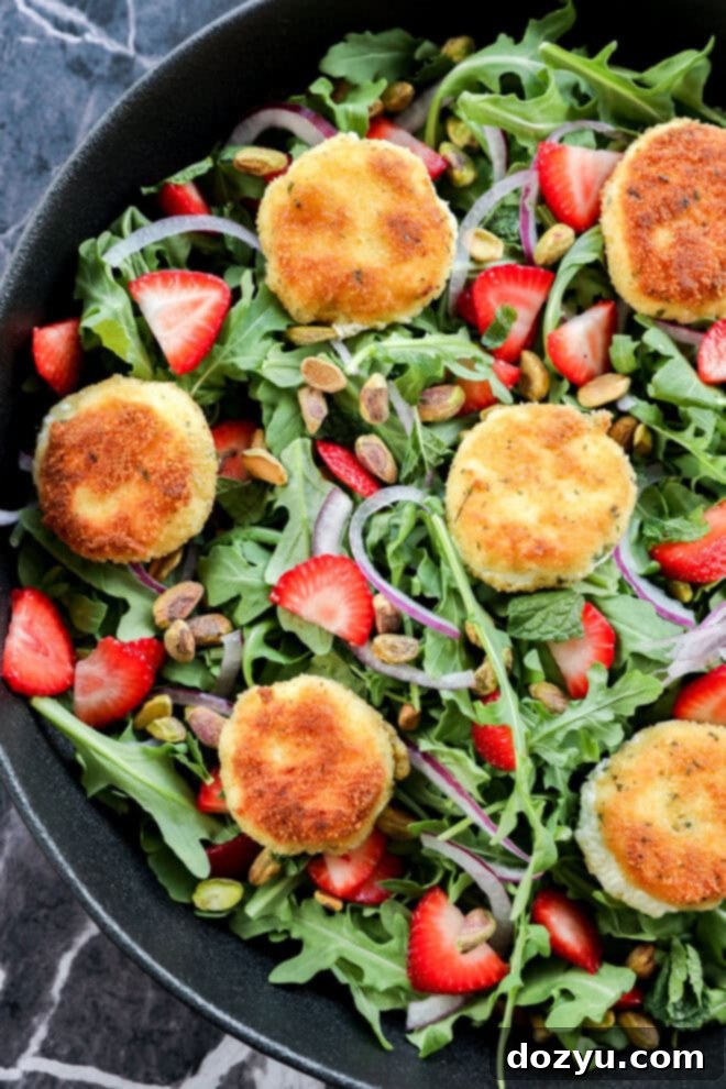 A salad with arugula, sliced strawberries, red onion, pistachios, and several breaded, golden-brown rounds of fried goat cheese, arranged in a black bowl on a dark marble surface.