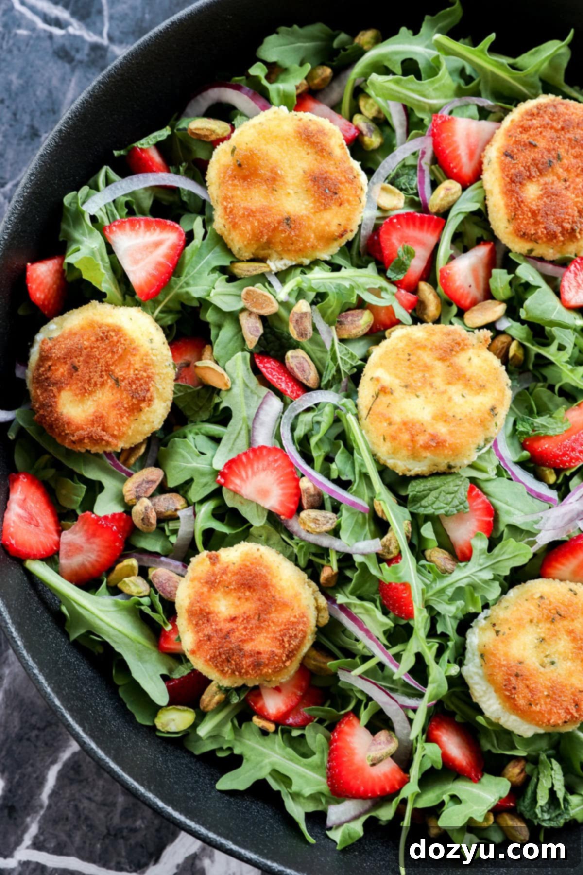 A salad with arugula, sliced strawberries, red onion, pistachios, and several breaded, golden-brown rounds of fried goat cheese, arranged in a black bowl on a dark marble surface.