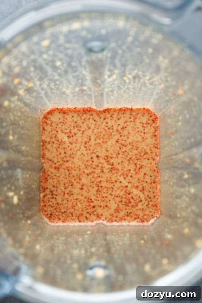 A close-up top view of a blender containing a tan liquid with small red specks, likely a blended sauce or dressing, with remnants clinging to the sides of the container.