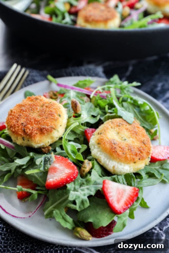 A plate of fresh arugula salad topped with sliced strawberries, roasted pistachios, and two golden-brown rounds of breaded goat cheese. A fork is beside the plate and more salad can be seen in the background.