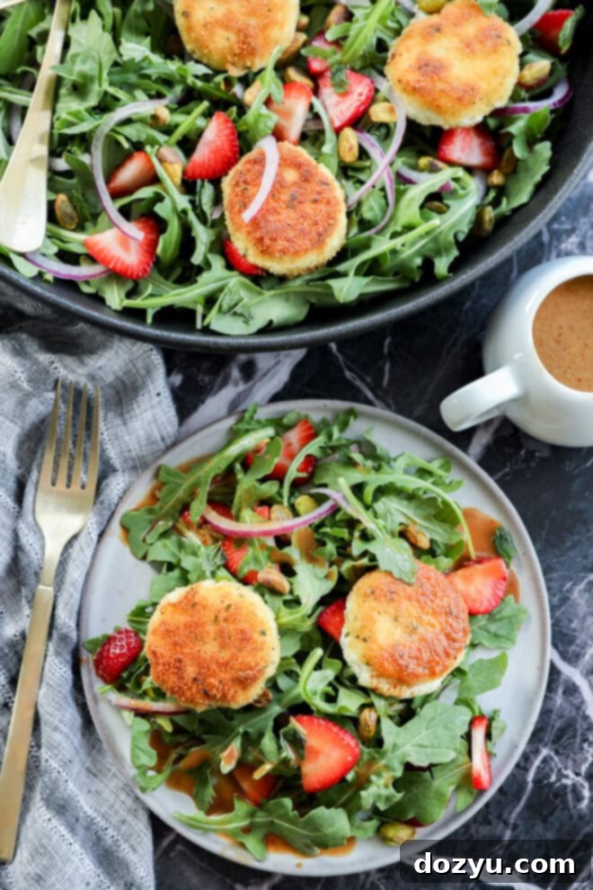 A fresh salad with arugula, sliced strawberries, red onions, pistachios, and crispy goat cheese rounds on a plate, with a fork, napkin, and small cup of dressing nearby.