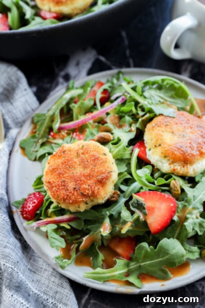 A plate of fresh arugula salad with sliced strawberries, red onions, pistachios, and two golden-brown breaded goat cheese rounds, drizzled with dressing.