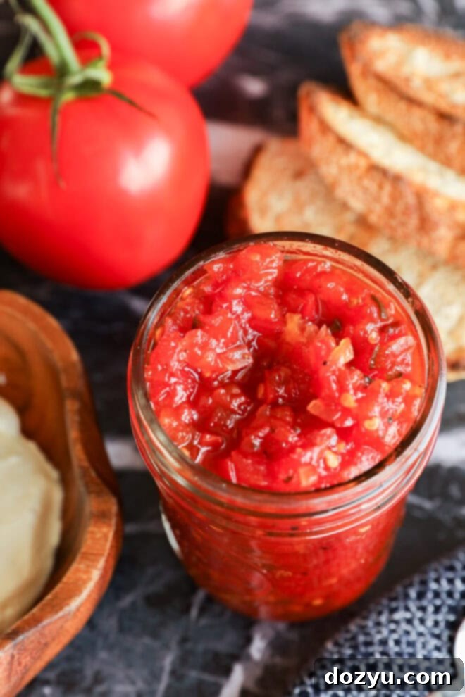 Artisan Tomato Relish 2 A glass jar filled with chunky tomato jam and tangy tomato relish sits on a table next to sliced bread, fresh tomatoes, and a wooden dish of butter.