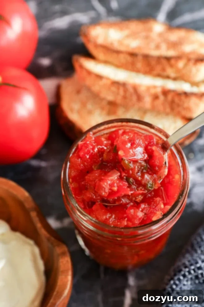 Artisan Tomato Relish 6 A jar of chunky tomato relish jam with a spoon inside, placed on a dark surface next to slices of toasted bread, fresh tomatoes, and a wooden bowl of cream cheese.