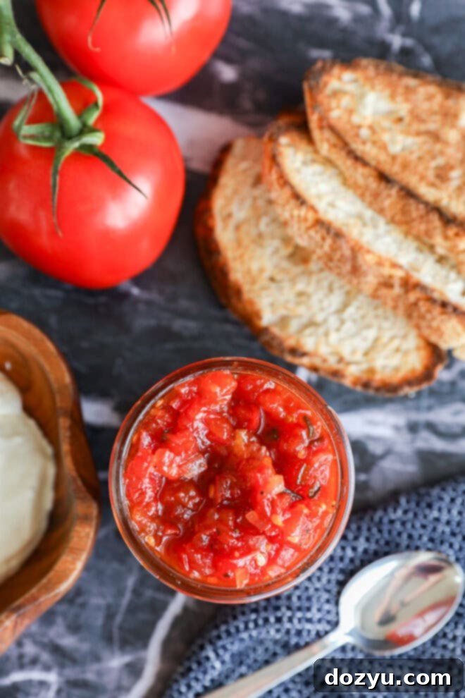 Artisan Tomato Relish 7 A jar of tomato relish sits on a marble surface next to slices of toasted bread, two fresh tomatoes on the vine, a bowl of white cheese, and a silver spoon.