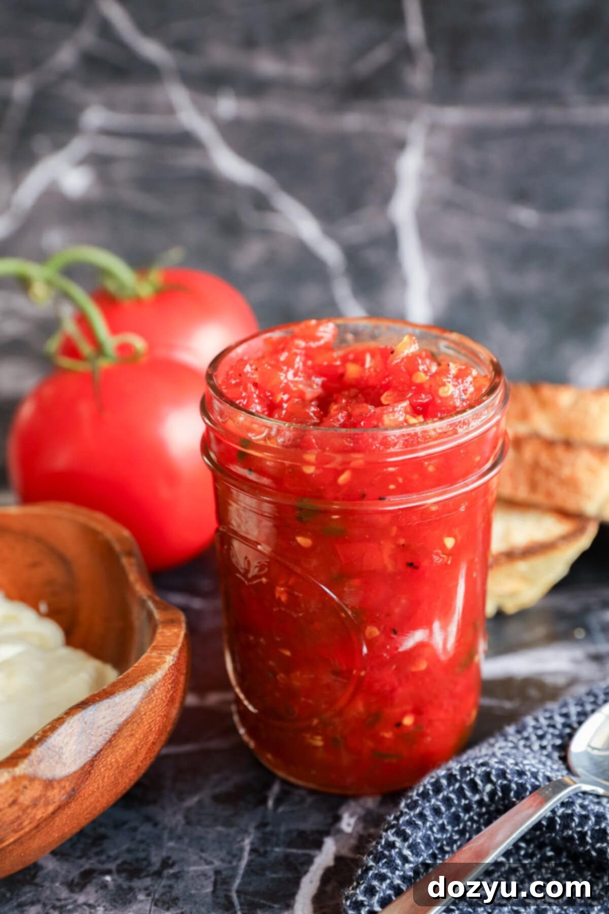 A glass jar filled with chunky tomato relish sits on a marble surface, surrounded by fresh tomatoes, slices of toasted bread, and a wooden bowl of butter.