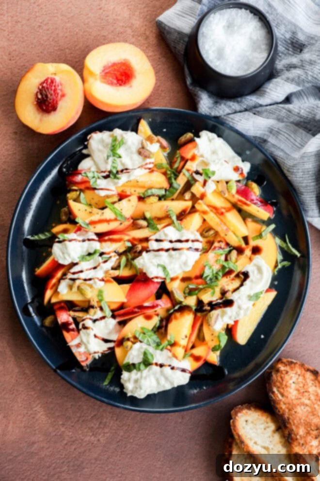 A plate of fresh salad with sliced peaches, burrata cheese, basil, and a drizzle of balsamic glaze, served with toasted bread. A halved peach and a bowl of salt are nearby on a brown surface.