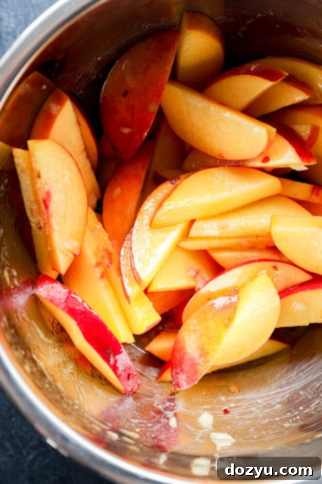 Sliced fresh peaches with red and yellow skin in a metal mixing bowl, lightly coated with a glaze or dressing.
