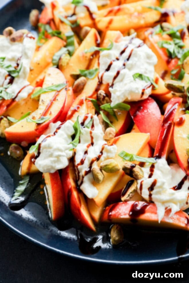 A close-up of a fresh salad with sliced peaches and nectarines, dollops of creamy cheese, chopped basil, pistachios, and drizzles of balsamic glaze, served on a dark plate.