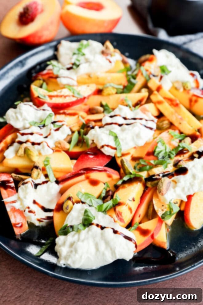 A plate of sliced peaches, fresh mozzarella or burrata, chopped pistachios, fresh basil, and a drizzle of balsamic glaze, with halved peaches in the background.