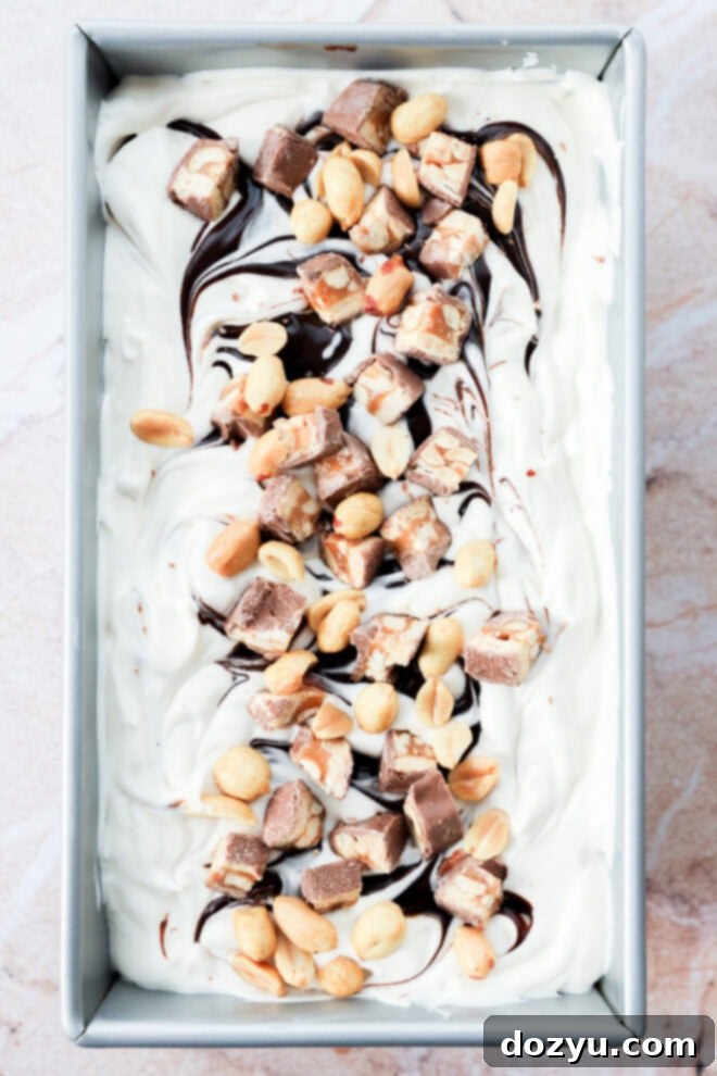 A rectangular pan filled with swirled vanilla dessert topped with chopped chocolate candy bars, peanuts, and ribbons of chocolate syrup.
