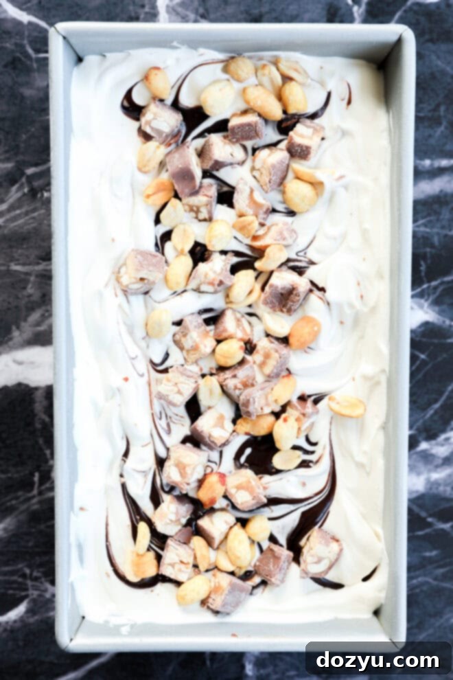 A rectangular pan filled with swirled vanilla dessert, topped with chocolate sauce, chopped candy bars, and peanuts, set on a dark marble surface.